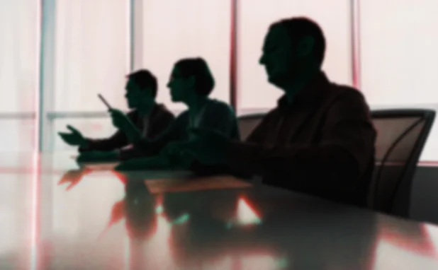 Blurred silhouettes of three businesspeople sat at a desk in an office, the middle person holding up a pen