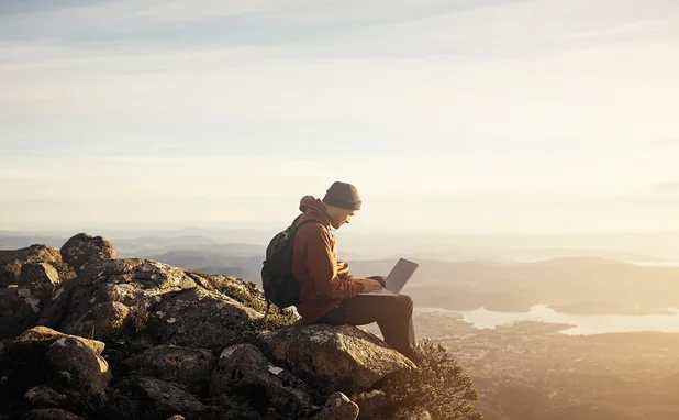 Man sits on a rock typing on a laptop, overlooking expansive countryside and river