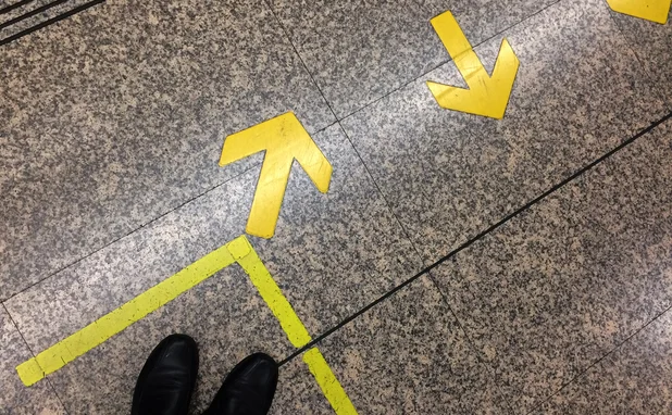 Top view of passenger’s feet standing in front of painted yellow box and directional arrows on a train station floor