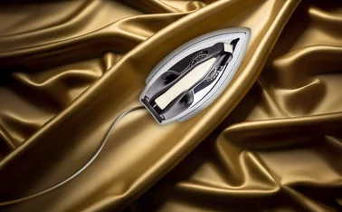 Vintage electric iron resting on luxurious gold satin fabric, highlighting the contrast between smooth textile folds and a sleek metal appliance