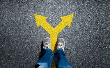 A person stands on a road, with a yellow arrow pointing in two different ways at their feet. 