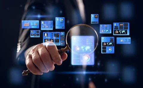 A man holds a magnifying glass that is magnifying one in a series of computer screens showing generic data