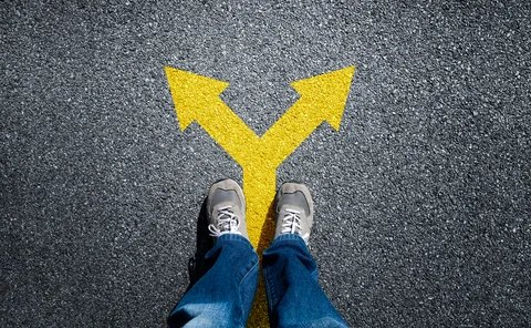 A person stands on a road, with a yellow arrow pointing in two different ways at their feet. 