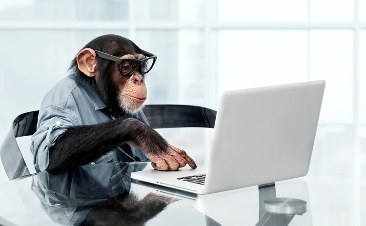 A chimpanzee in spectacles and a blue shirt is sitting by a laptop.  