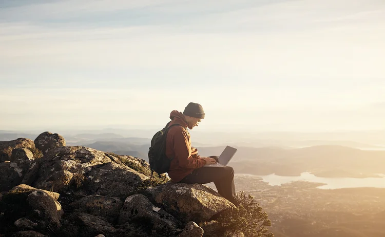 Man sits on a rock typing on a laptop, overlooking expansive countryside and river