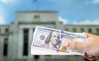 A hand holding US dollars in front of the Federal Reserve