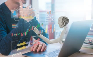 Man sitting in front of laptop, while a virtual robot leans out of the screen and looks at financial data 