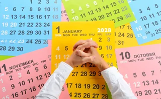 Several calendar pages are strewn on a table, with a person’s arms resting on the table with clasped hands, showing a decision must be made