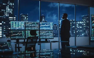 Businessman stands in front of large window, contemplating multiple trading screens at an empty desk at night 