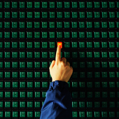 A finger switches on a button, which lights up red, on a panel full of green buttons that are switched off