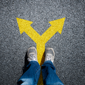 A person stands on a road, with a yellow arrow pointing in two different ways at their feet. 