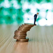 Businessman figurine standing on stack of coins that looks like it's about to topple over